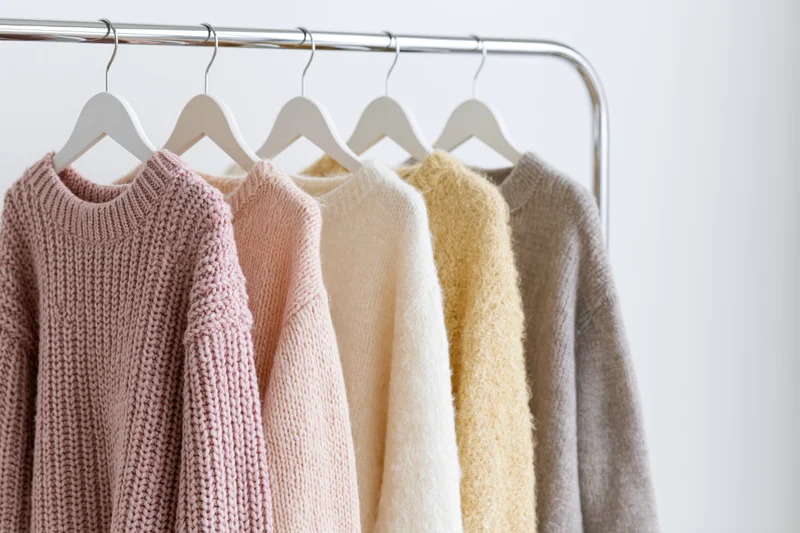 Five knit sweaters in pastel shades hanging on white hangers along a metal rack against a plain white wall