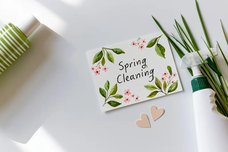 Handwritten spring cleaning card with floral decoration on white surface surrounded by cleaning bottles and greenery