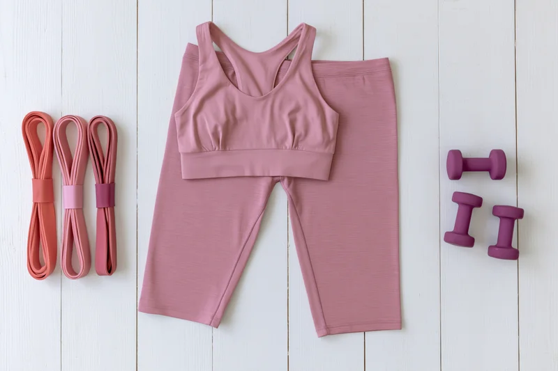 Pink sports bra and matching leggings with resistance bands and dumbbells arranged on white wooden surface