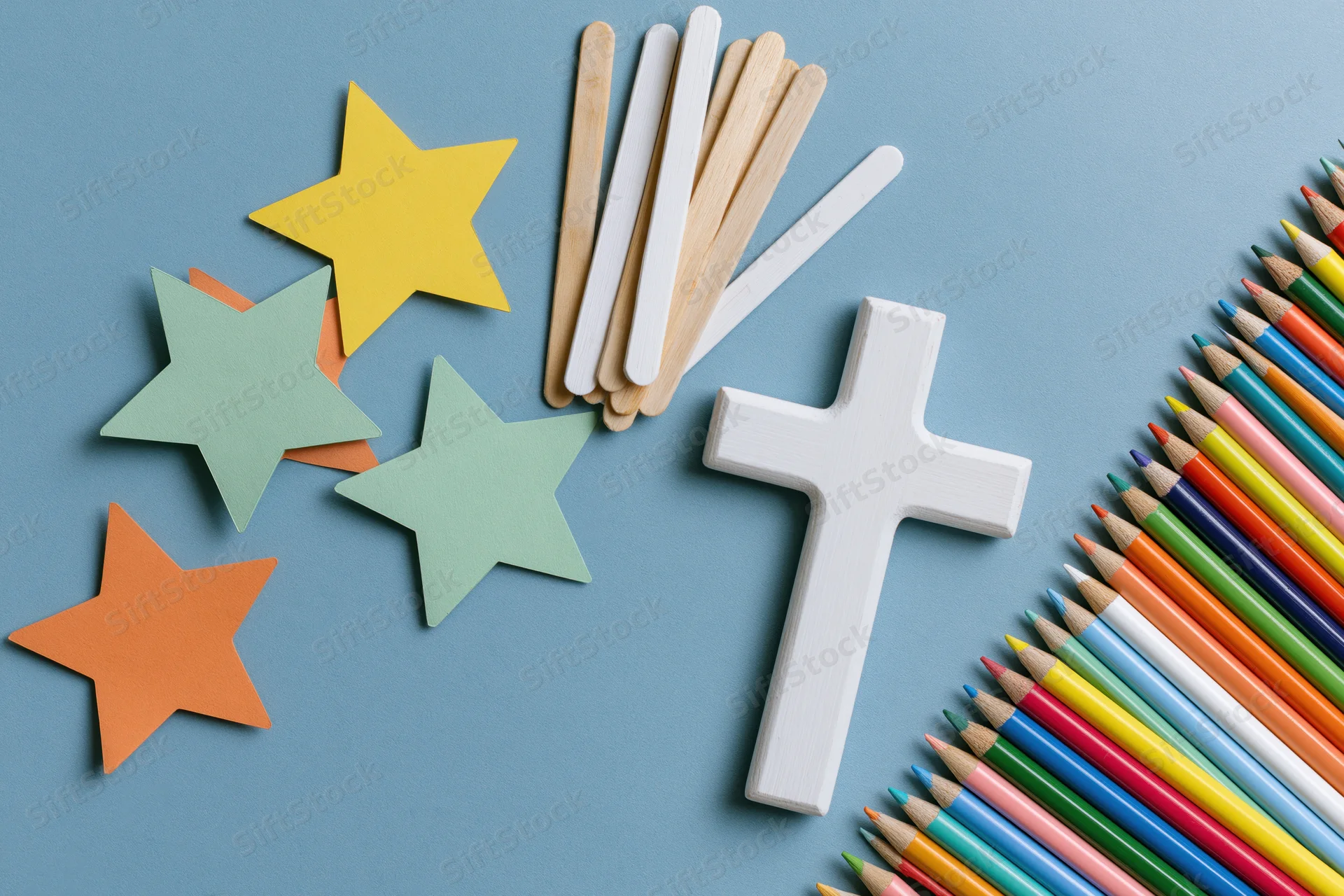 White wooden christian cross with colorful craft sticks and paper stars arranged on blue background