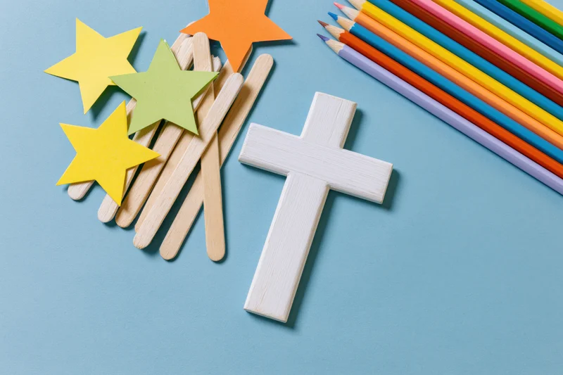 White wooden christian cross with craft sticks and colored pencils on blue background