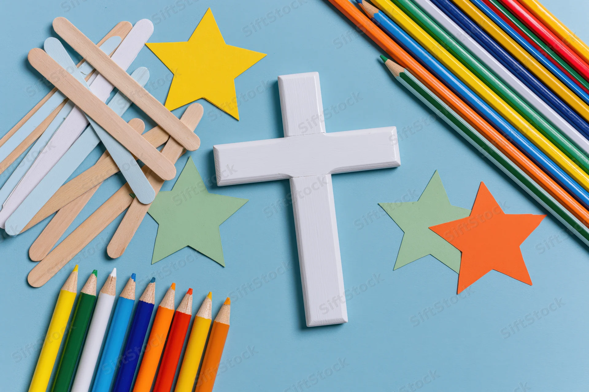 White wooden christian cross surrounded by craft sticks, colored pencils and paper stars on blue surface