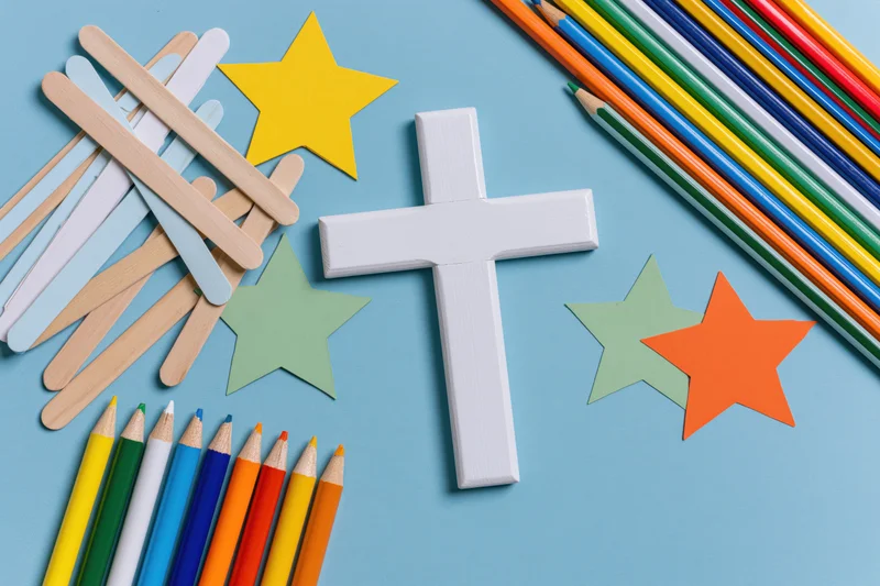 White wooden christian cross surrounded by craft sticks, colored pencils and paper stars on blue surface