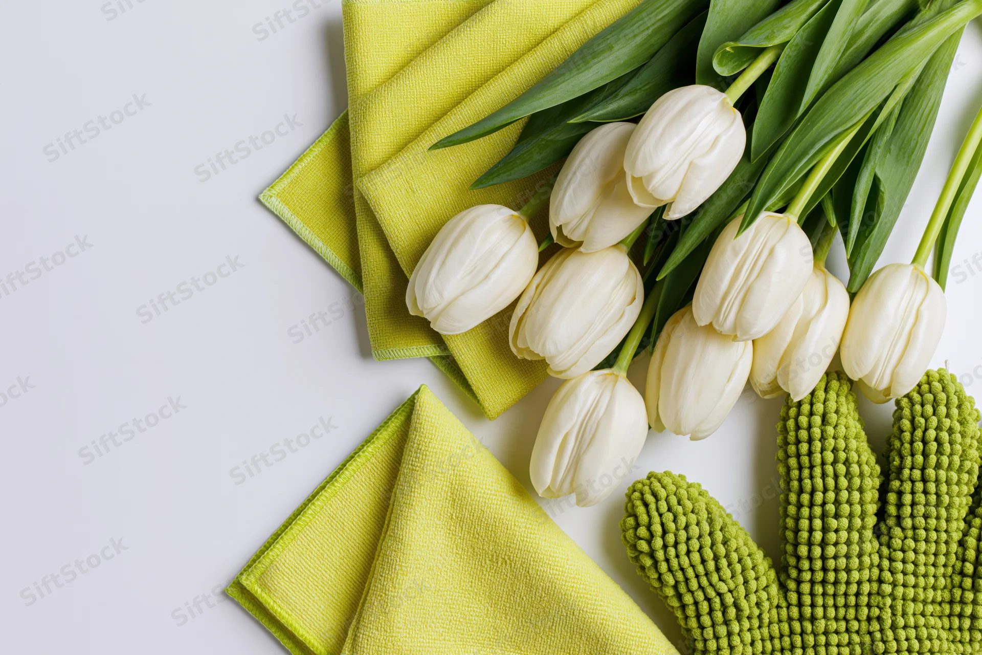Cream tulips with green leaves arranged alongside yellow napkins and green gloves on white surface