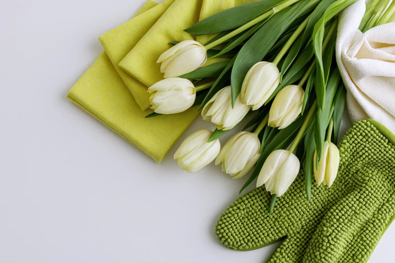 Cream tulips with green leaves arranged alongside folded napkins and gardening gloves on a white surface