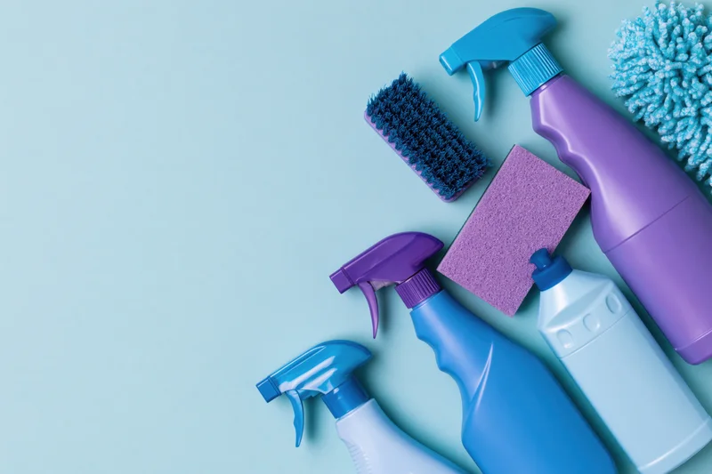 Household cleaning supplies arranged in flat lay on light blue surface with copyspace