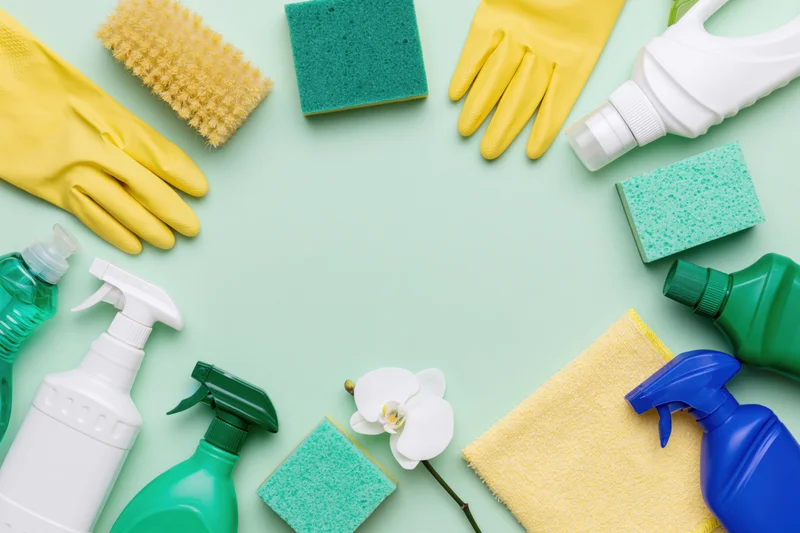 Household cleaning supplies arranged in a loose circular border on a light green surface with copyspace in center