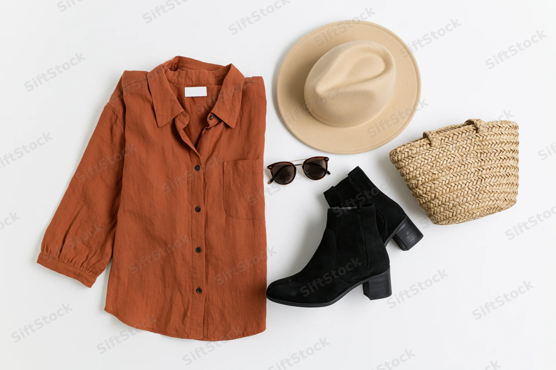 Flat lay of women's autumn outfit with rust shirt, hat, boots, sunglasses and woven bag on white surface