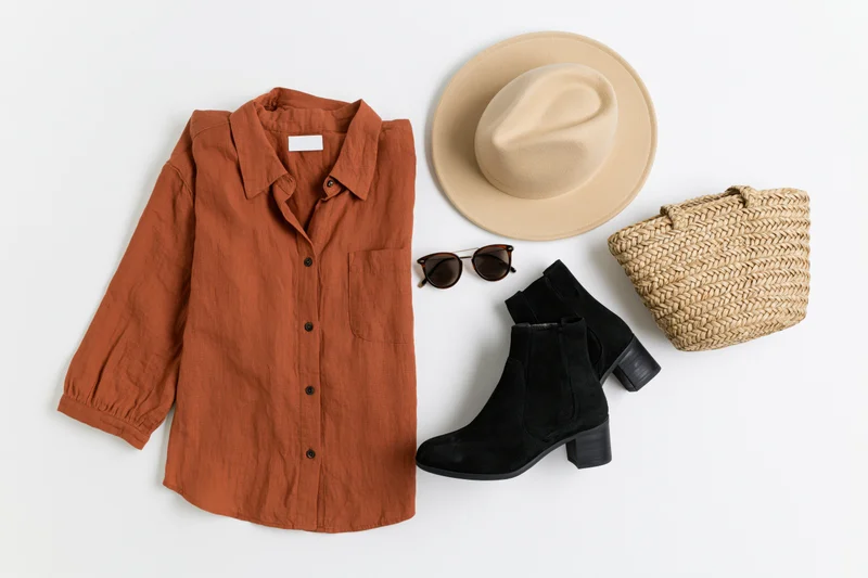Flat lay of women's autumn outfit with rust shirt, hat, boots, sunglasses and woven bag on white surface