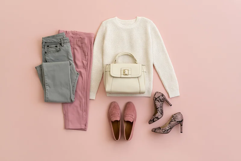 Women's fashion flat lay with sweater, jeans, trousers, handbag, loafers and heels on pink background