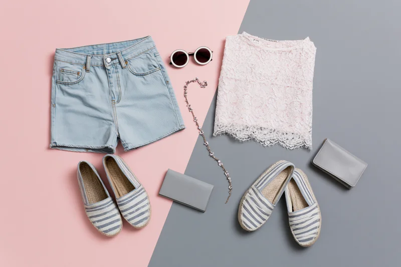 Women's summer outfit with denim shorts, lace top, espadrilles, sunglasses, wallet and necklace arranged on pink and gray background