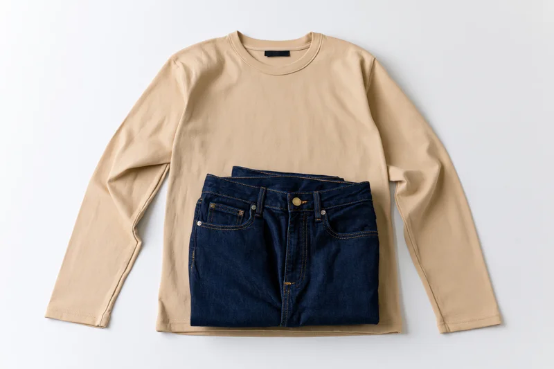 Beige long-sleeve t-shirt folded with dark blue jeans stacked on top, flat lay on white background