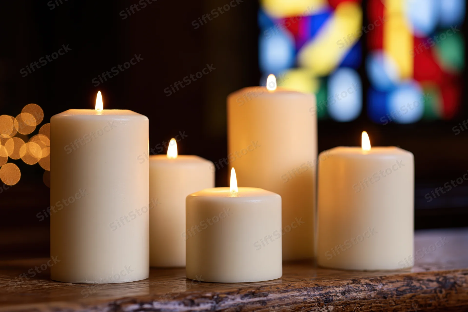 Lit white pillar candles arranged on a wooden table in front of stained glass window in church interior