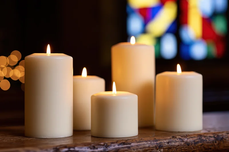 Lit white pillar candles arranged on a wooden table in front of stained glass window in church interior