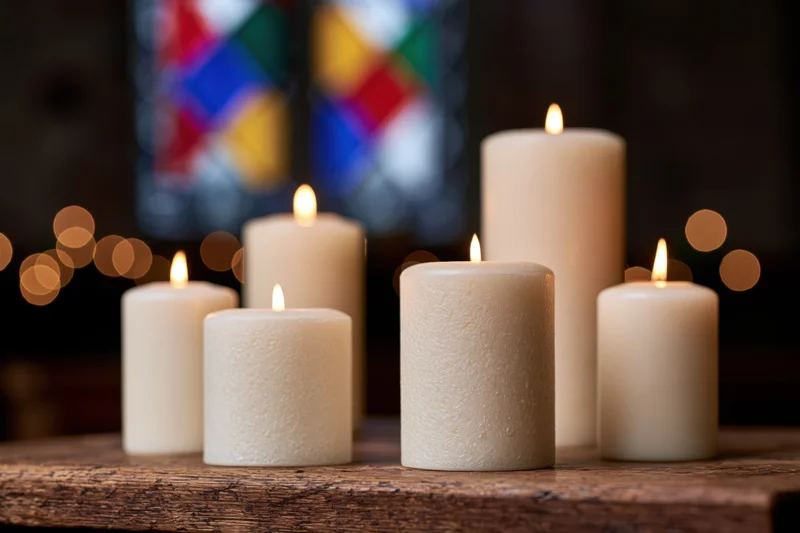 Lit white pillar candles of varying heights arranged on a wooden table in front of stained glass window