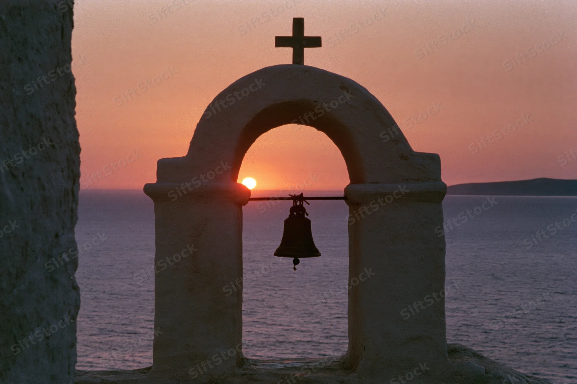 Bell in stone arch of church bell tower silhouetted against sunset over sea