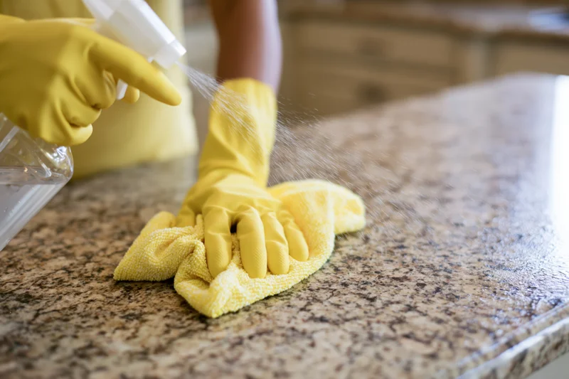 Hands in yellow rubber gloves spraying cleaning liquid onto a granite kitchen countertop while holding a cloth