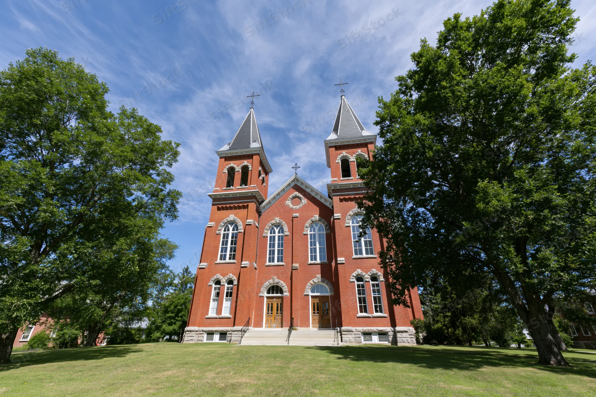 Red brick catholic church with twin spires and arched windows on a grassy lawn under a blue sky