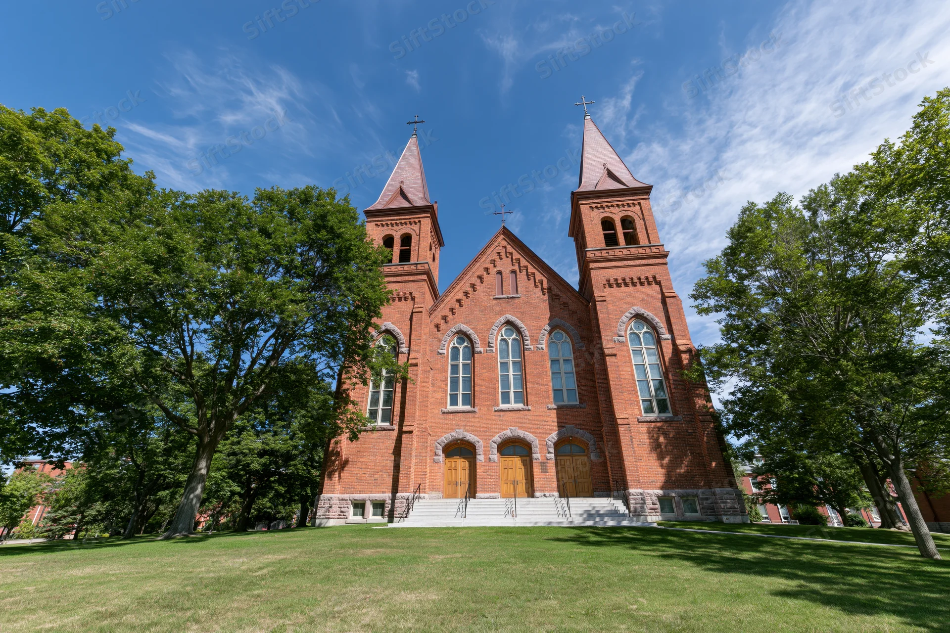 Historic red brick catholic church with twin spires and arched windows on a grassy lawn under a blue sky