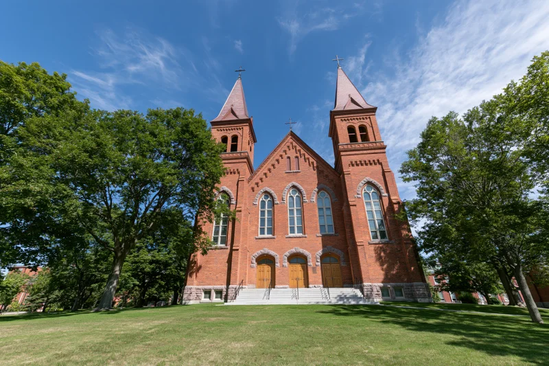 Historic red brick catholic church with twin spires and arched windows on a grassy lawn under a blue sky
