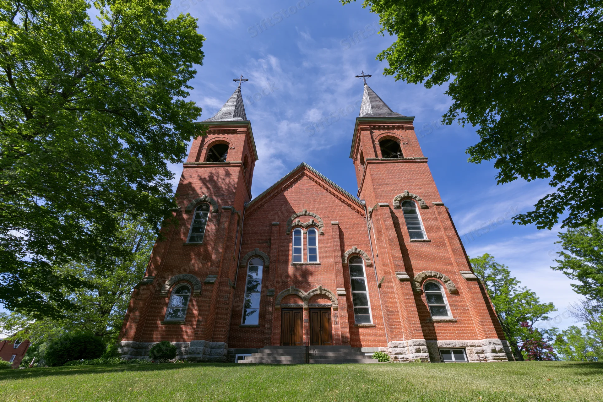 Red brick catholic church with twin spires and arched windows on a grassy lawn under blue sky