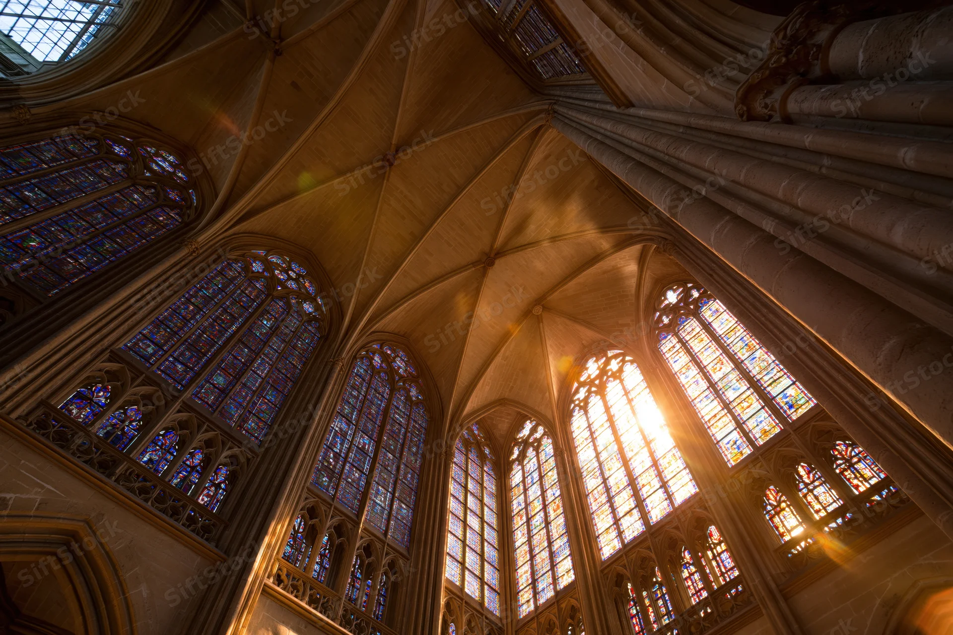 Upward view of gothic cathedral nave interior with vaulted stone ceiling and stained glass windows illuminated by sunlight