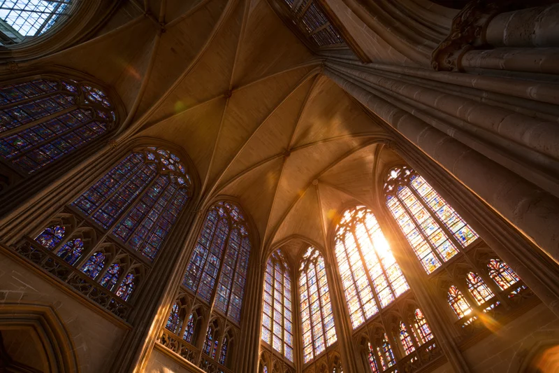 Upward view of gothic cathedral nave interior with vaulted stone ceiling and stained glass windows illuminated by sunlight