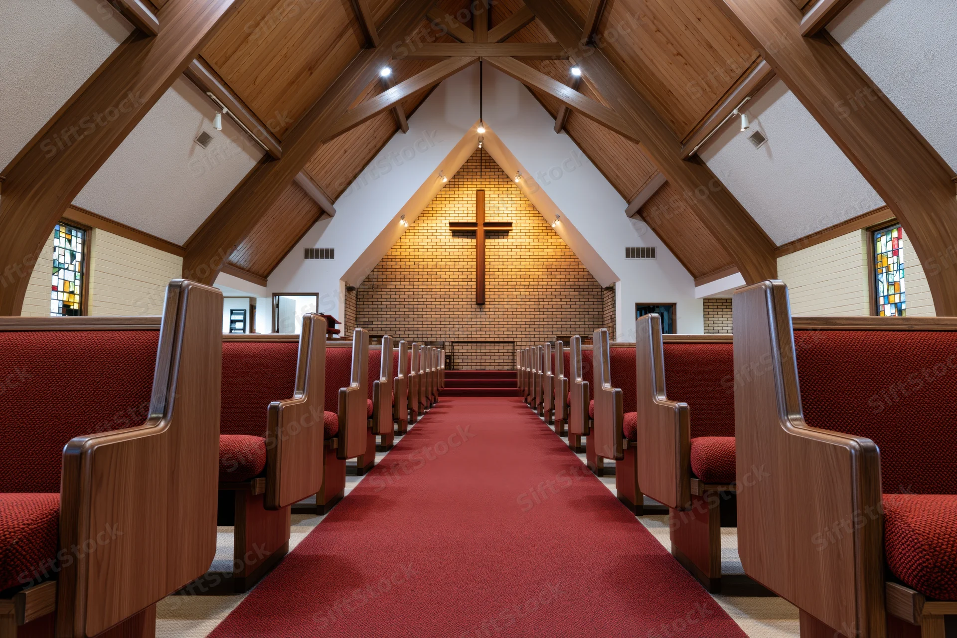 Empty wooden pews with red upholstery lining a central aisle in a modest protestant church nave with a brick altar and cross under a vaulted wooden ceiling