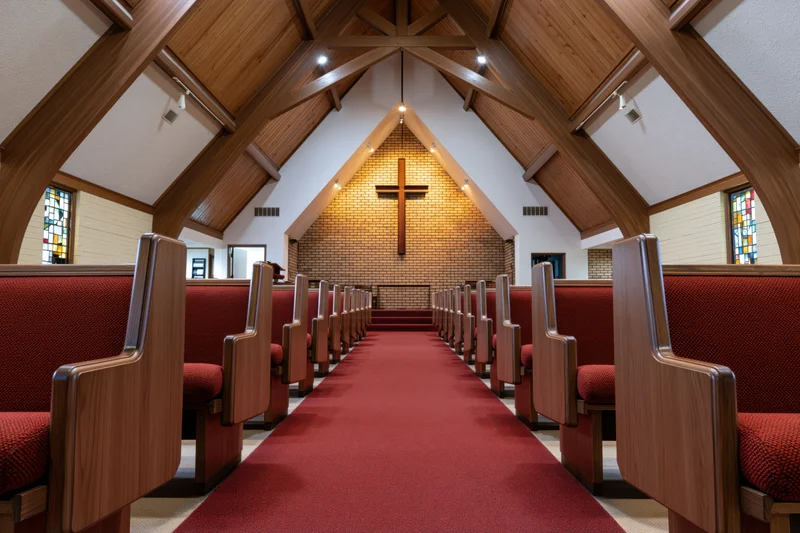 Empty wooden pews with red upholstery lining a central aisle in a modest protestant church nave with a brick altar and cross under a vaulted wooden ceiling