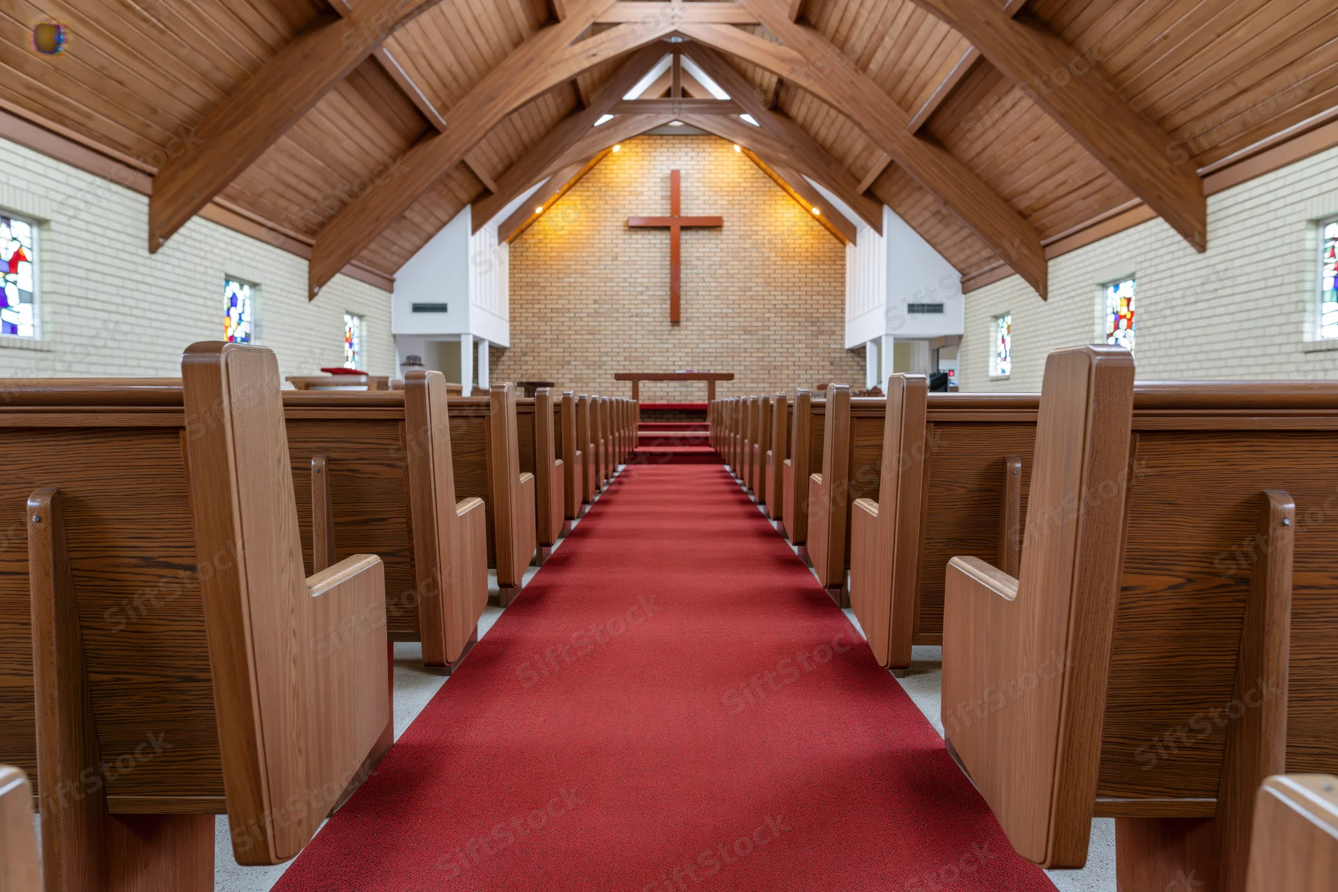 Empty wooden pews lining a red carpeted aisle leading to a cross on a brick altar in a modest protestant church interior