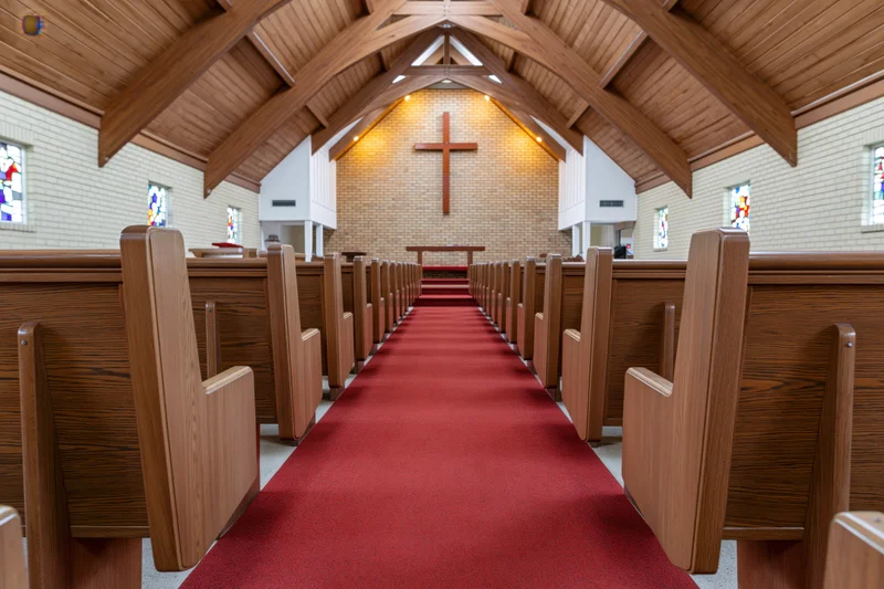 Empty wooden pews lining a red carpeted aisle leading to a cross on a brick altar in a modest protestant church interior