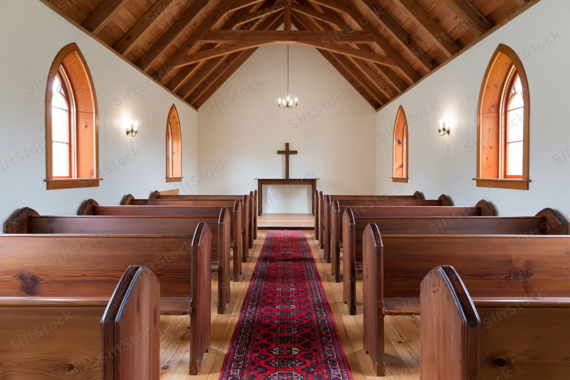 Empty wooden pews lining a central aisle with a red rug leading to an altar and cross in a small chapel interior