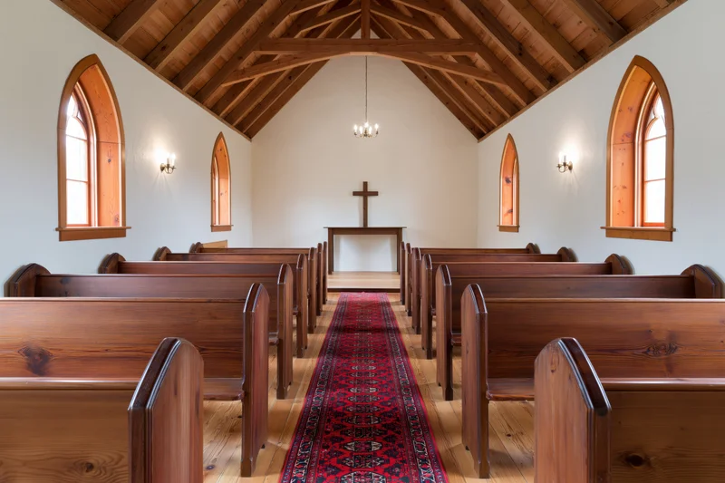 Empty wooden pews lining a central aisle with a red rug leading to an altar and cross in a small chapel interior