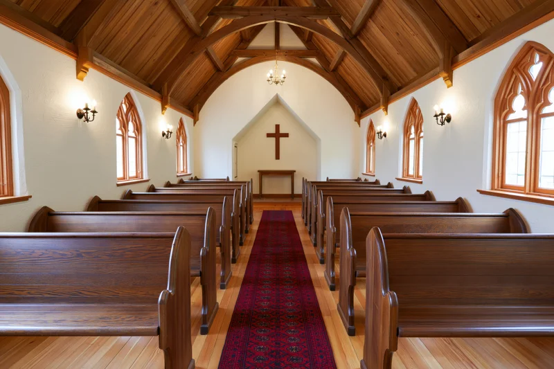 Wooden chapel interior with empty pews lining a red rug aisle toward a cross at the altar