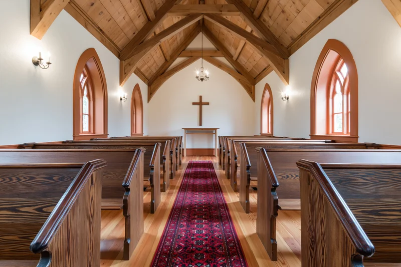 Empty wooden pews lining a central aisle with a red rug leading to an altar and cross in a small chapel interior