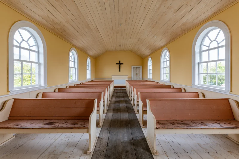Empty wooden pews arranged in rows inside a small historic church with yellow walls and arched windows