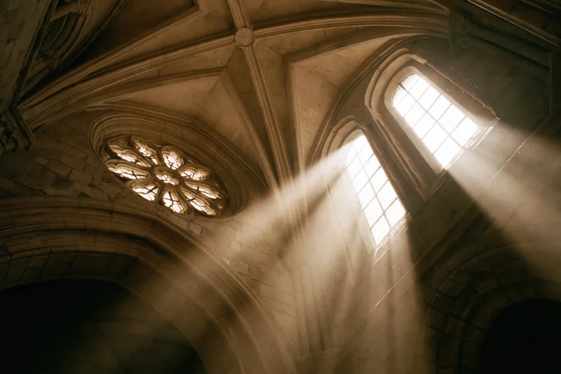 Interior of a stone gothic church with sunlight streaming through arched windows and a rose window in the vaulted ceiling