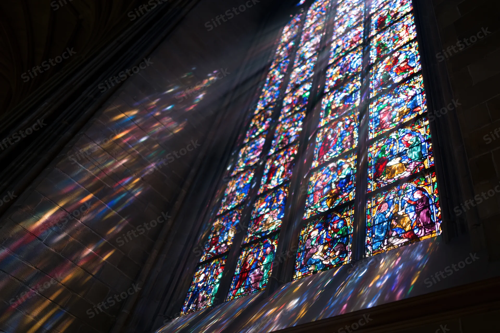 Large gothic stained glass window in cathedral interior with sunlight streaming through and colorful reflections on stone walls and ceiling