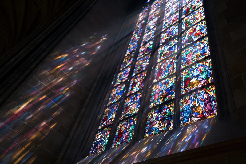 Large gothic stained glass window in cathedral interior with sunlight streaming through and colorful reflections on stone walls and ceiling