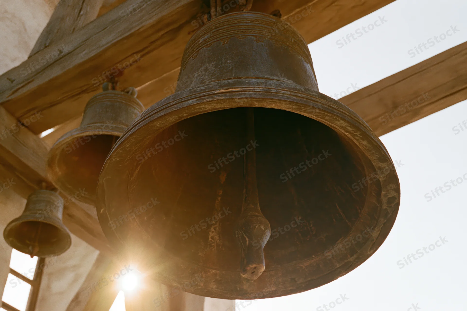 Large aged bronze church bell hanging from wooden beam in belfry with sunlight shining through