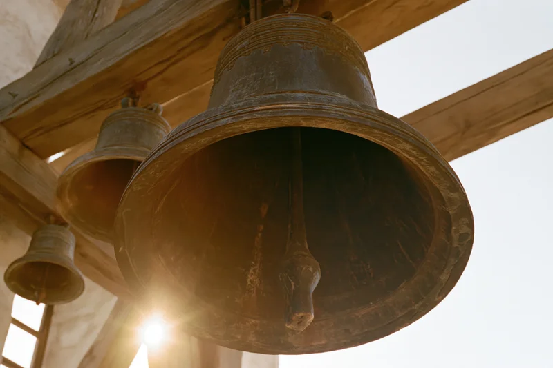 Large aged bronze church bell hanging from wooden beam in belfry with sunlight shining through