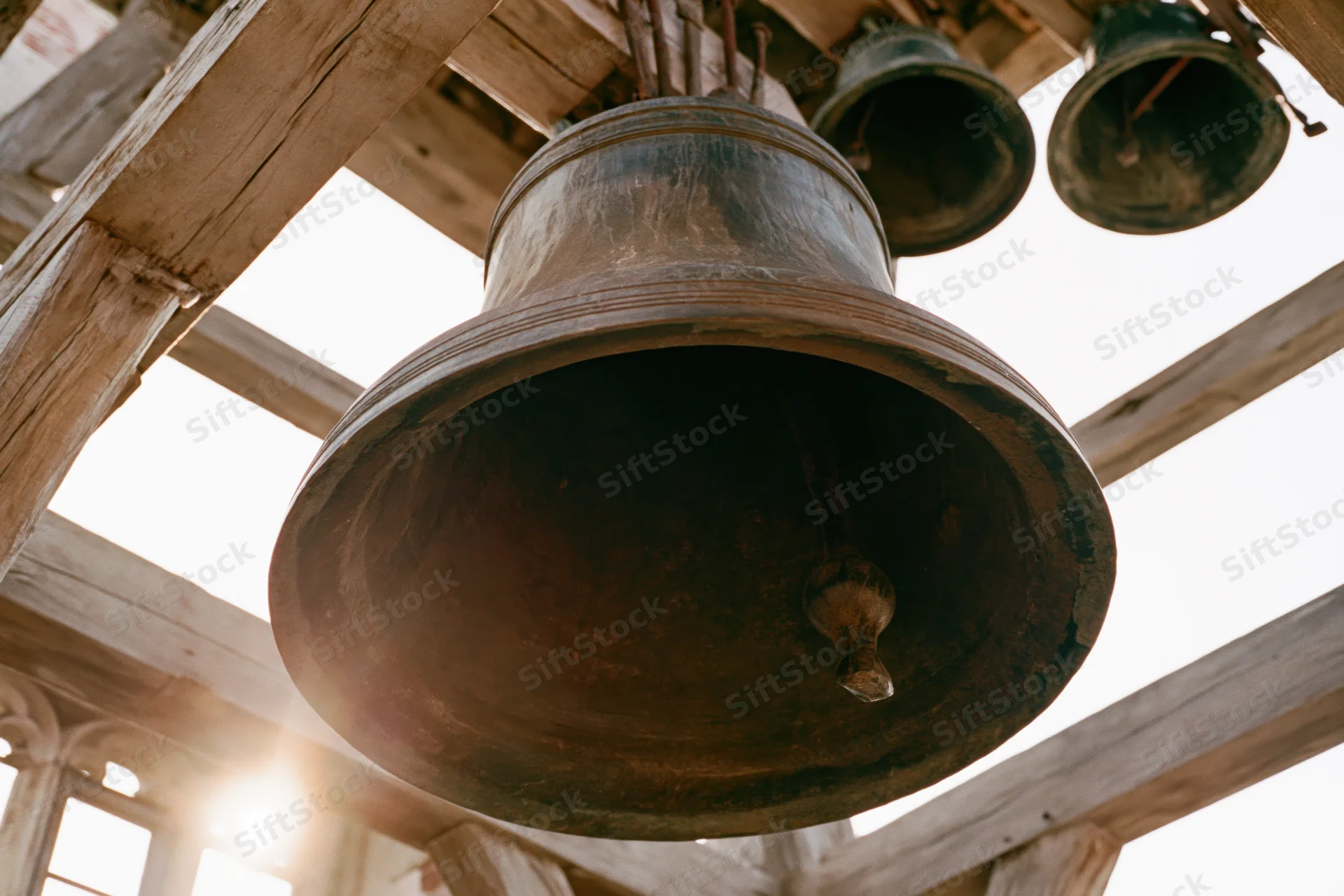 Large aged bronze church bell hanging from wooden beams in a belfry with sunlight flaring through
