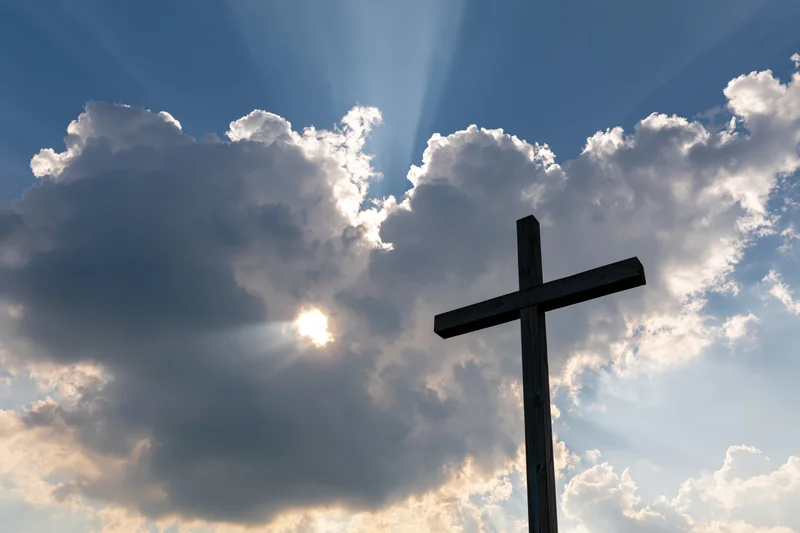 Wooden christian cross silhouetted against a dramatic sky with sunbeams breaking through clouds
