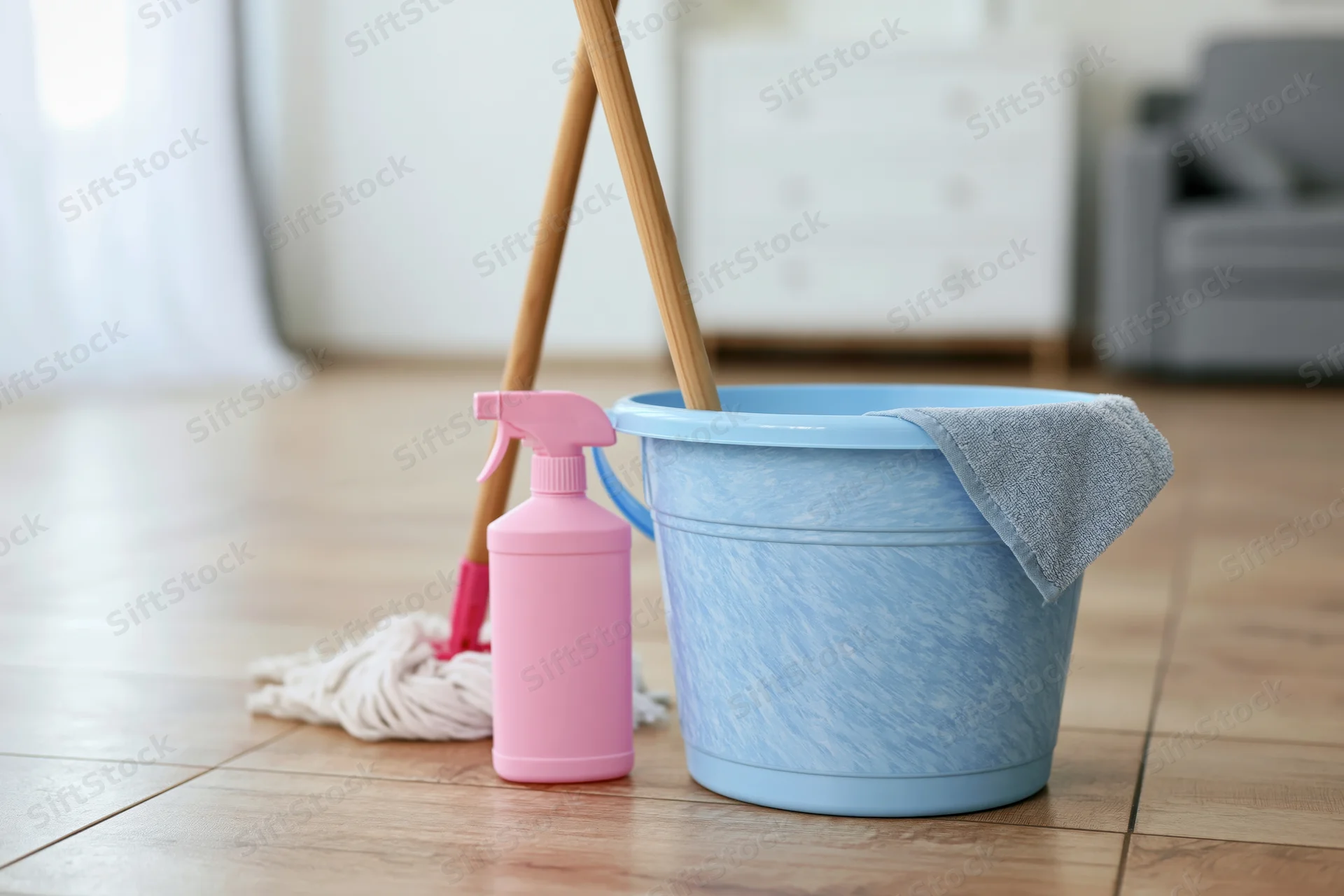 Light blue plastic bucket with mop and pink spray bottle on wooden floor in living room