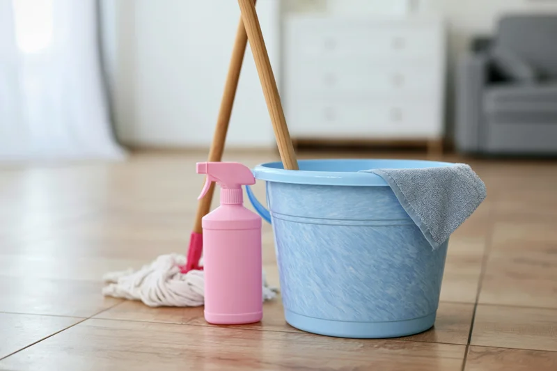 Light blue plastic bucket with mop and pink spray bottle on wooden floor in living room