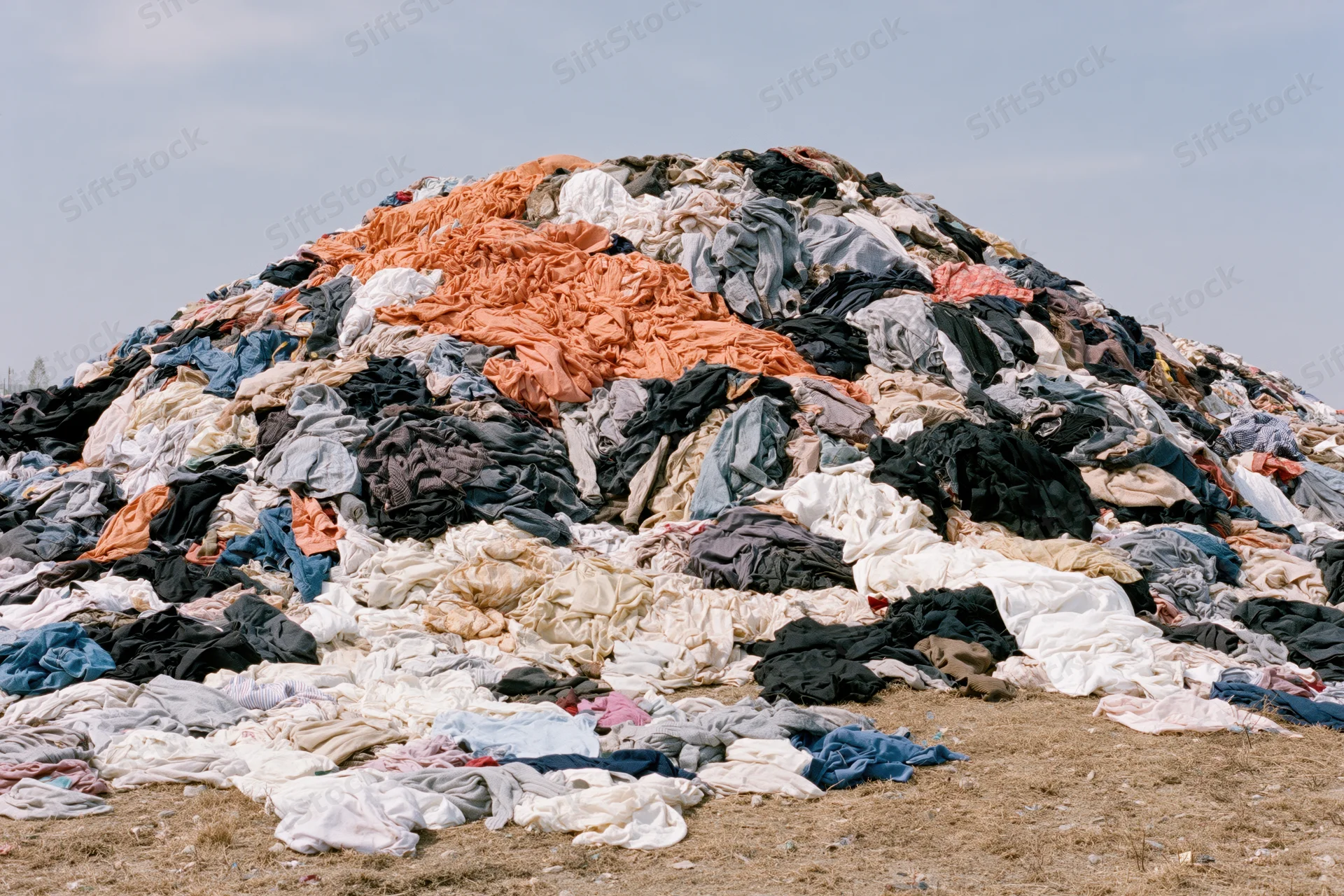 Massive mound of discarded clothing piled high outdoors on dry ground under a pale sky