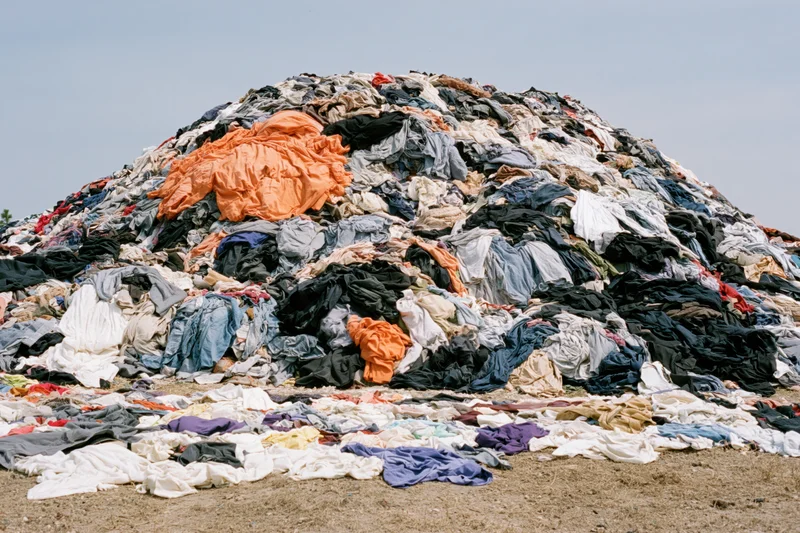 Massive mound of discarded clothing piled high outdoors on dry ground under clear sky