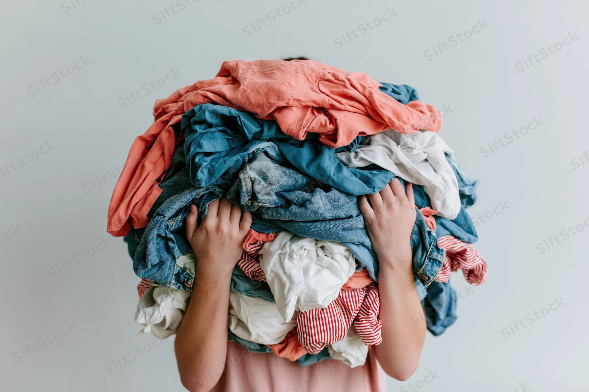 Woman holding large pile of laundry covering her face in a plain indoor setting