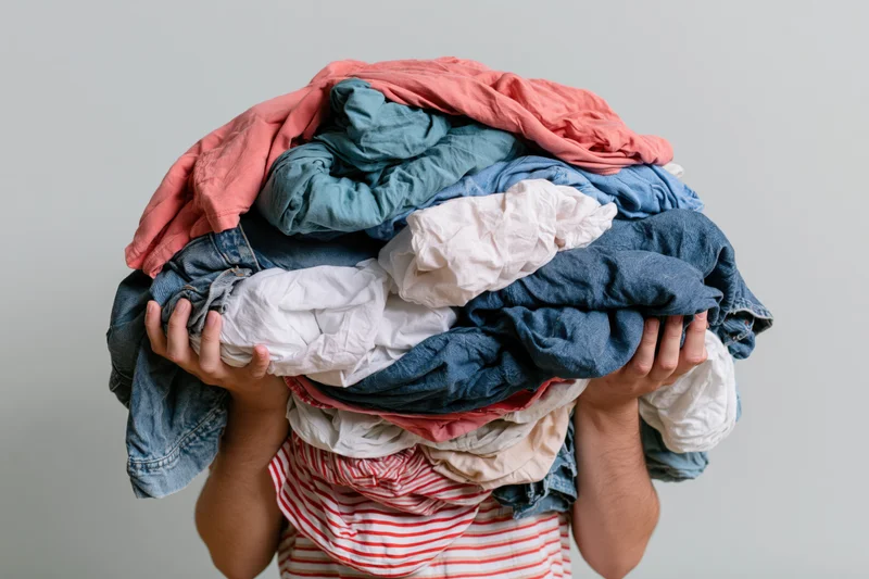 Man holding large pile of laundry over his head, obscuring his face, against plain background