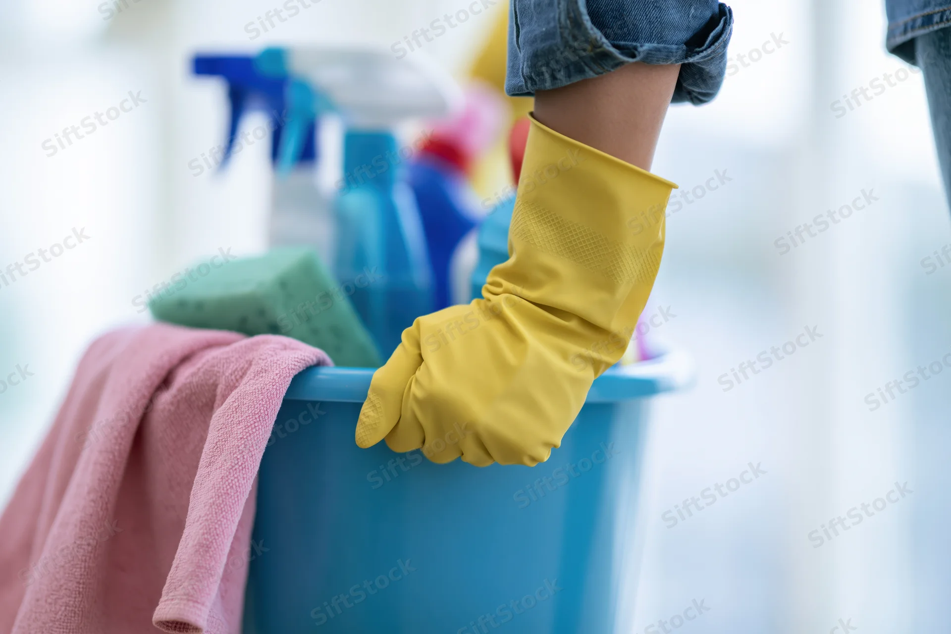 Woman's hand in yellow rubber glove holding blue plastic bucket filled with cleaning supplies and towel